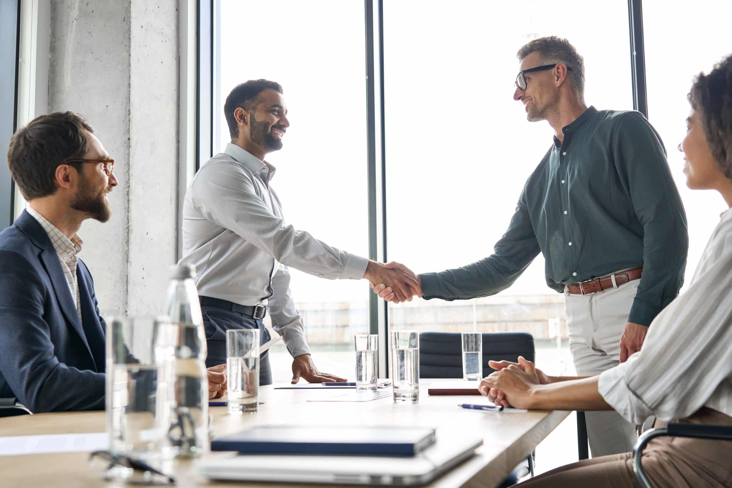 Two businessmen shake hands across a modern conference table, symbolizing a new partnership agreement.